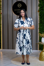 A woman stands in a boutique wearing a white short-sleeve midi dress with a navy blue floral and bird pattern. The background features vertical wood slats, a black circular sign reading 'AYANU BOUTIQUE', and a green wall with a gold side table.