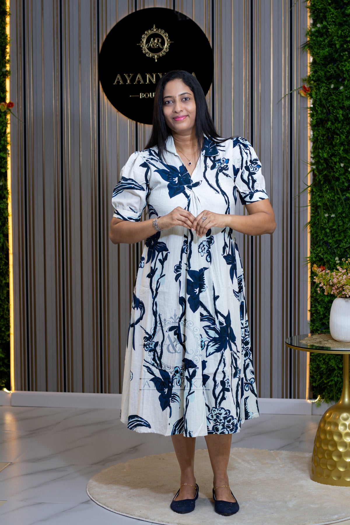 A woman stands in a boutique wearing a white short-sleeve midi dress with a navy blue floral and bird pattern. The background features vertical wood slats, a black circular sign reading 'AYANU BOUTIQUE', and a green wall with a gold side table.