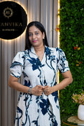 A woman wearing a white short-sleeve dress with a navy blue floral print stands in front of a decorative wall with a green hedge and a vertical striped panel. A black circular sign on the left reads 'ANVIKA BOUTIQUE'.