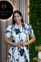 A woman wearing a white short-sleeve dress with a navy blue floral print stands in front of a decorative wall with a green hedge and a vertical striped panel. A black circular sign on the left reads 'ANVIKA BOUTIQUE'.