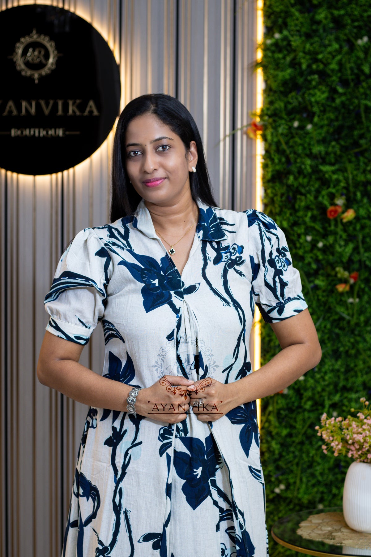 A woman wearing a white short-sleeve dress with a navy blue floral print stands in front of a decorative wall with a green hedge and a vertical striped panel. A black circular sign on the left reads 'ANVIKA BOUTIQUE'.
