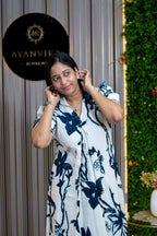 A woman posing in a white short-sleeve button-up shirt with a navy blue floral print, set against a modern background featuring a vertical green wall and a black circular sign.