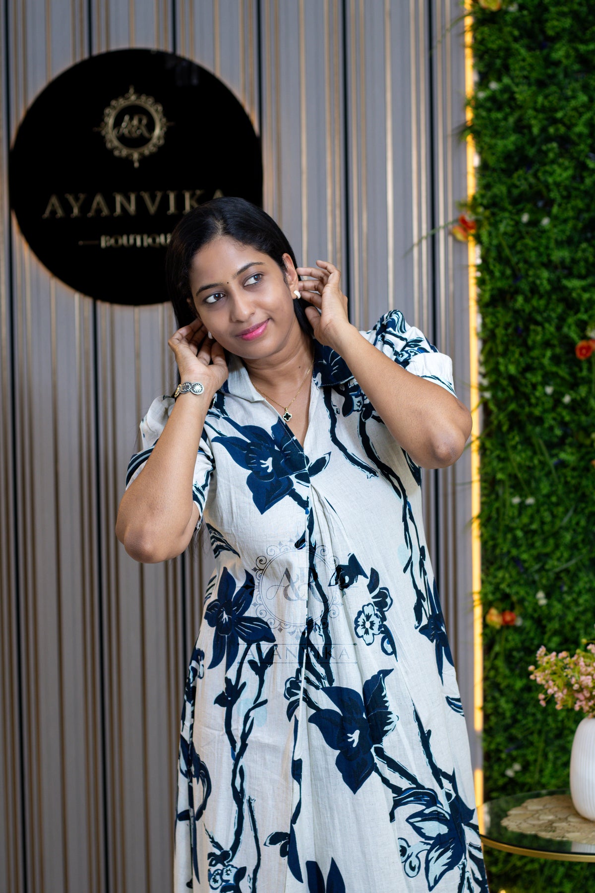 A woman posing in a white short-sleeve button-up shirt with a navy blue floral print, set against a modern background featuring a vertical green wall and a black circular sign.