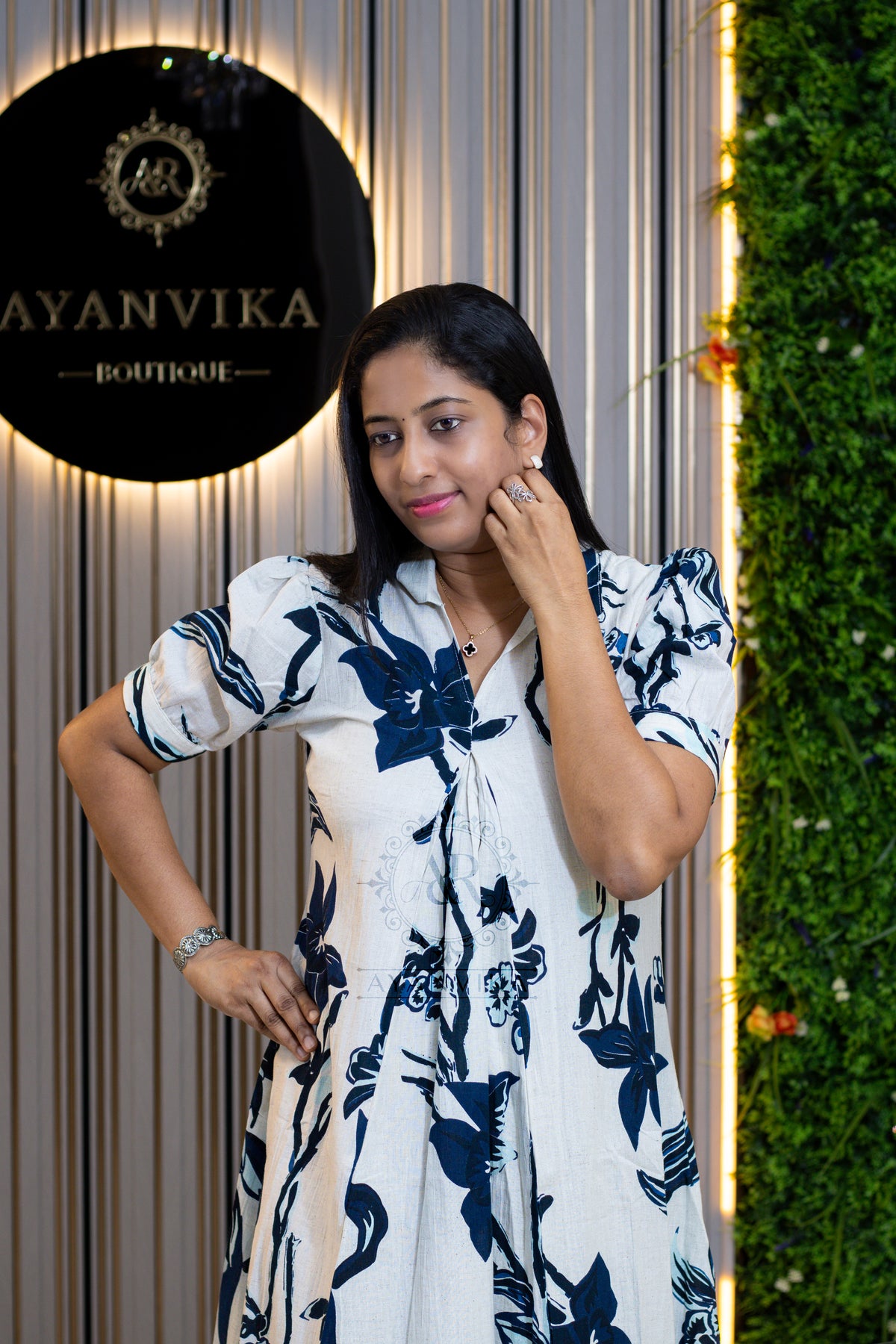 A woman posing in a white short-sleeve button-up dress with a navy blue floral pattern, standing in front of a boutique backdrop with a circular 'AYANVIKA BOUTIQUE' sign and a green wall.